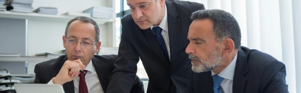 Drei Geschäftsmänner in Anzügen betrachten konzentriert einen Laptop-Bildschirm in einem modernen Büro. Das Bild veranschaulicht die zunehmende Relevanz der FlexCo Österreich in der Rechtsanwaltsbranche.