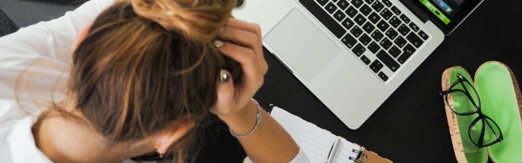 Eine gestresste Frau mit den Händen im Haar sitzt vor einem Laptop auf einem schwarzen Tisch. Das Bild illustriert die typischen Fehler und Herausforderungen bei der Einrichtung einer flexiblen Kapitalgesellschaft.