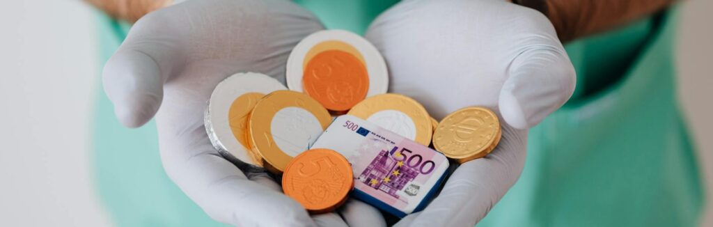 Arzt in grüner Kleidung hält Euro-Münzen und Geldschein aus Schokolade – symbolisiert die steuerlichen Grundlagen und Finanzen für Ärzte in Österreich.