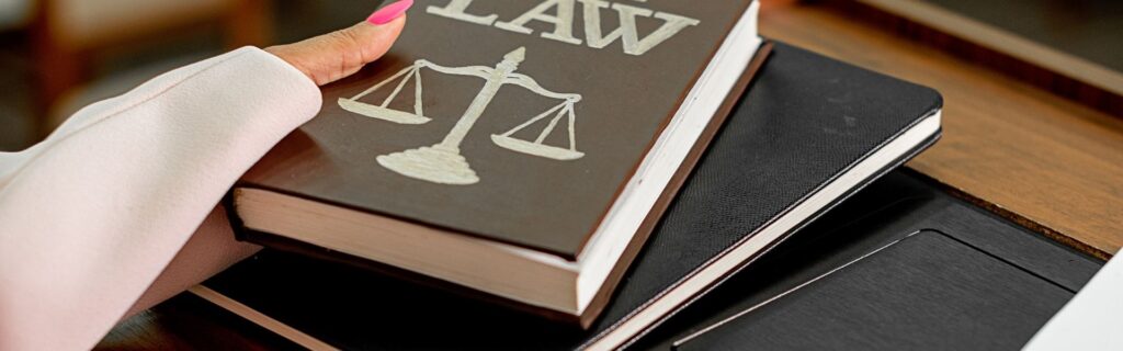 Hand hält ein Gesetzbuch mit Justitia-Symbol und dem Schriftzug „LAW“, daneben liegen Notizbücher auf einem Schreibtisch. Das Bild steht für rechtliche Änderungen und neue steuerliche Vorschriften im Budgetbegleitgesetz 2025.