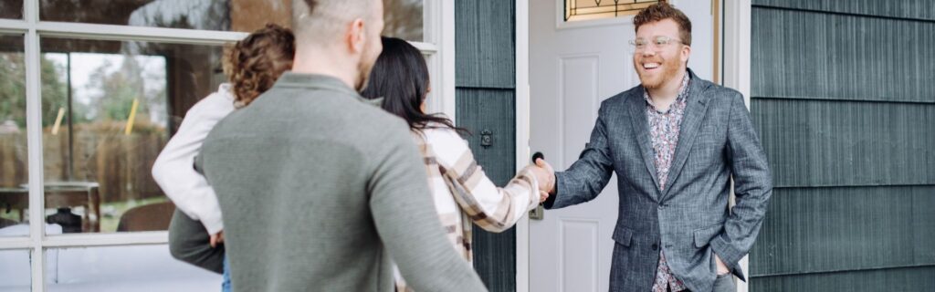 Zwei Personen geben sich vor dem Eingang eines Hauses die Hand, während eine Familie im Hintergrund steht. Das Bild symbolisiert Immobilienkauf, Investorenentscheidungen und die Auswirkungen der neuen Share Deal-Regeln.