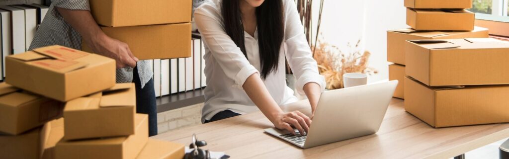 Junge Unternehmerin arbeitet am Laptop und ist umgeben von Versandkartons – steht für typische Herausforderungen und Praxisbeispiele von KMU und Start-ups bei der Zinsschranke 2026.