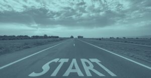 Eine gerade Landstraße erstreckt sich unter wolkigem Himmel bis zum Horizont, am vorderen Bildrand ist groß das Wort "START" auf den Asphalt gemalt. Die Umgebung ist ländlich mit Feldern und wenigen Bäumen zu beiden Seiten der Straße. Das Titlebild steht für den Beginn einer neuen Reise oder eines neuen Abschnitts – genau wie der Gründungsprozess eines Unternehmens einen bedeutsamen Neustart im Leben eines Unternehmers markiert.