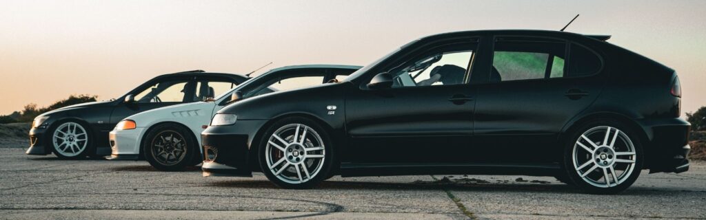 Alt: Drei Pkw im Seitenprofil nebeneinander auf leerem Parkplatz bei Sonnenuntergang; steht für den Kostenvergleich Firmenwagen 2025 – E‑Auto versus Diesel/Benziner – inklusive Unterschiede bei Sachbezug, motorbezogener Versicherungssteuer und jährlichen Gesamtkosten.