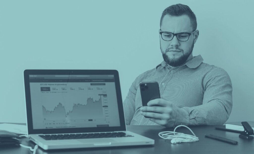 Abgebildet ist ein Geschäftsmann an einem modernen Schreibtisch mit Laptop, Smartphone und Arbeitsunterlagen. Im Hintergrund ist ein Börsengraph am Laptop zu sehen. Das Bild steht für unternehmerische Strategie, Jahresabschluss und Planung mit dem Steuerberater.