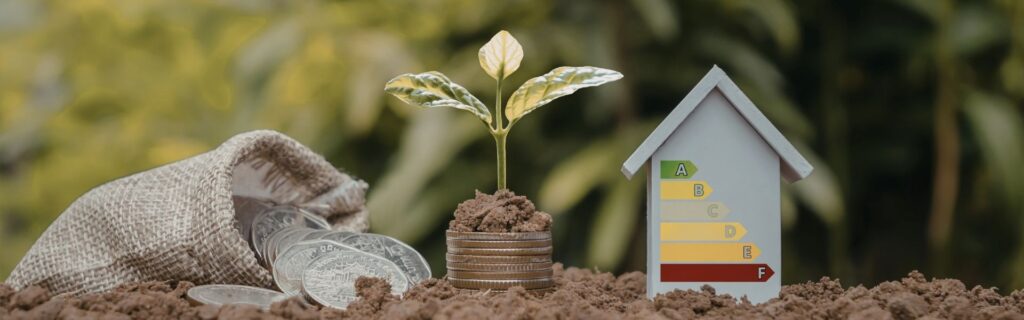Ein Säckchen mit silbernen Münzen liegt auf dem Erdboden, daneben ein gestapeltes Bündel Münzen mit wachsendem Pflänzchen; rechts ein Modellhaus mit Energie-Effizienzlabel von A bis F. Das Bild steht für die neuen steuerlichen Vorteile durch den erhöhten Investitionsfreibetrag 2025 und nachhaltige Anschaffungen. Investitionsfreibetrag ab November 2025 – Symbolfoto für Steueränderungen: gestapeltes Münzgeld mit grünem Pflänzchen sowie Energie-Effizienzhaus und Geldsack, Fokus auf nachhaltige Investitionen und Steuerersparnis für Unternehmen.