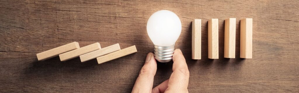 Investitionsfreibetrag optimal nutzen – Symbolbild. Mehrere Holz-Dominosteine kippen nach links, werden jedoch von einer Hand gestoppt, die auch eine leuchtende Glühbirne hält. Rechts stehen noch weitere Dominosteine. Das Bild symbolisiert intelligente Planung und den richtigen Einsatz des Investitionsfreibetrags für eine sichere steuerliche Zukunft.