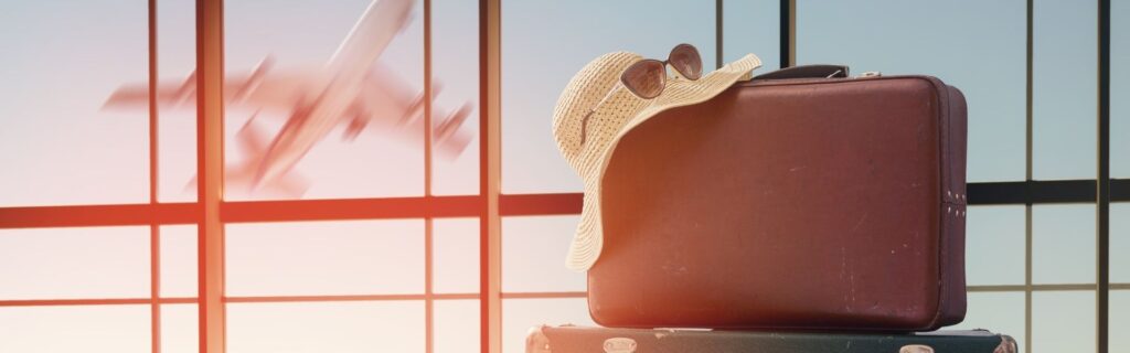 Brauner Koffer mit Sonnenhut und Sonnenbrille steht vor einem großen Fenster im Flughafen, ein Flugzeug ist im Hintergrund unscharf erkennbar; symbolisiert Anspruch und Planung von BUAK-Urlaub und Urlaubsgeld für Bauarbeiter in Österreich.