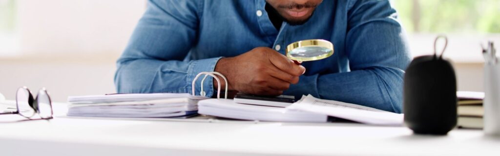 Mann prüft im Büro mit einer Lupe akribisch seine Dokumente; steht für die exakte Kontrolle des Jahressechstels und die strategische Vermeidung von Steuerüberhang bei Weihnachtsgeld und Bonuszahlungen 2025.