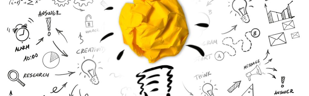 Gelbes Papierknäuel als Glühbirne vor Skizzen von Ideen und Strategien, symbolisch für den Weg von der ersten Business-Idee zur ersten Rechnung