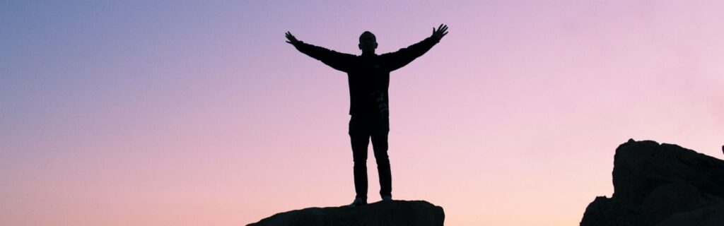 Schwarze Silhouette einer Person, die mit weit ausgebreiteten Armen auf einem Felsen oder erhöhten Punkt steht. Der Hintergrund zeigt einen dramatischen Sonnenuntergang mit rosa-violettem Himmel. Die Körperhaltung drückt Triumph, Freiheit und Erfolg aus. Das Bild symbolisiert den Erfolgsmoment von Immigrant-Gründern, die ihre Ziele erreicht haben – für den Abschnitt über erfolgreiche Branchen für migrantische Unternehmer in Österreich.
