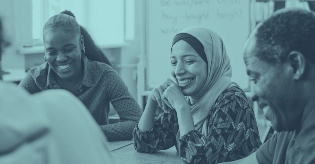 Drei professionell gekleidete Geschäftsfrauen unterschiedlicher Herkunft sitzen an einem modernen Konferenztisch in Wien. Eine Frau mit Kopftuch lächelt herzlich während eines Geschäftsgesprächs. Das Bild hat einen türkis-blauen Überlagerungs-Filter und symbolisiert erfolgreiche migrantische Unternehmerinnen, die gemeinsam an Geschäftsideen arbeiten. Die Atmosphäre ist freundlich, professionell und inklusiv.