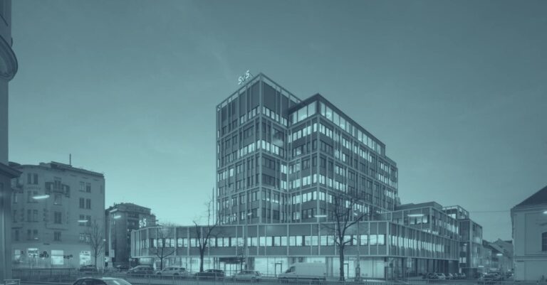 SVS-Beiträge 2026: Hauptgebäude der Sozialversicherung der Selbständigen (SVS) in Wien bei Dämmerung. Moderner neunstöckiger Bürokomplex mit Glasfassade und beleuchteten Fenstern in türkis-blauem Licht. SVS-Logo an Gebäudespitze. Symbolbild für SVS-Beitragsgrundlage, Sozialversicherung Selbständige und Gewinnfreibetrag-Optimierung 2026 in Österreich.