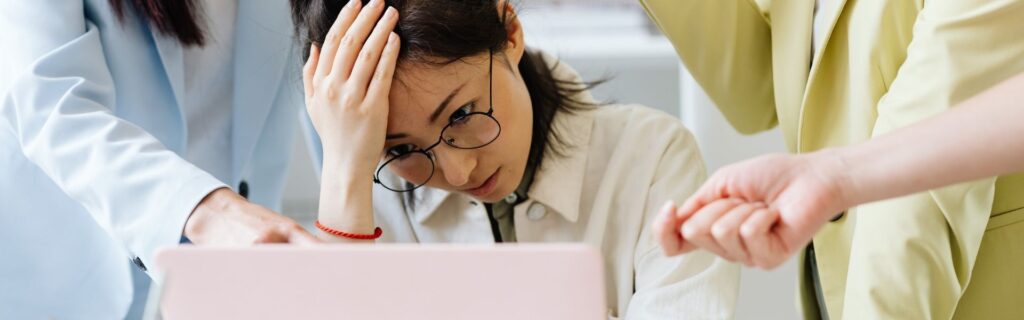 Unternehmerin sitzt gestresst vor dem Laptop, zwei Personen zeigen auf den Bildschirm – symbolisiert die Unsicherheit rund um das Doppelbudget