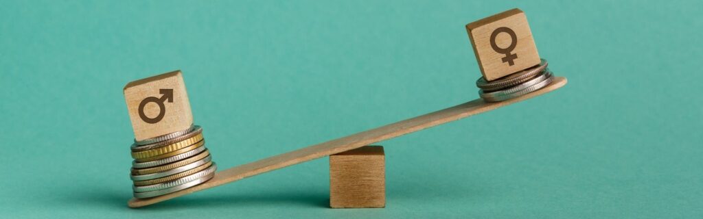 Waage aus Holz vor grünem Hintergrund, links ein Stapel Münzen mit männlichem Symbol, rechts weniger Münzen mit weiblichem Symbol. Deutlich sichtbare Schräglage als Darstellung des Gender Pay Gap und ungleicher Entlohnung von Frauen und Männern.