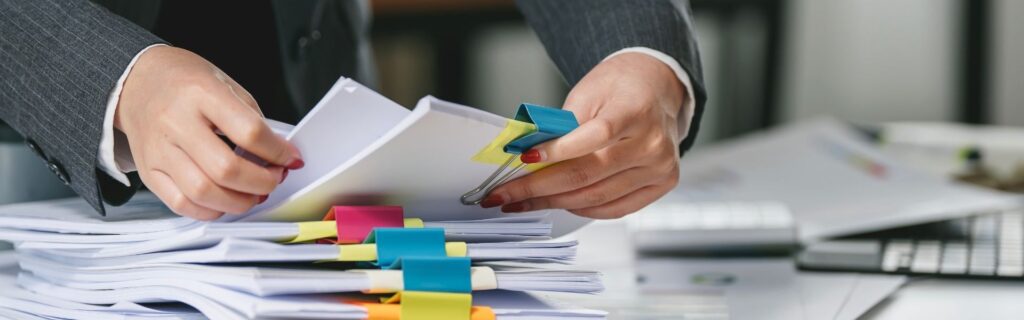 Geschäftsperson ordnet mehrere Papierstapel mit farbigen Büroklammern neben einem Laptop. Symbolbild für Entsendungen nach Österreich, ZKO3-Meldung, A1 Bescheinigung und vollständige Lohnunterlagen für DACH-Unternehmen.