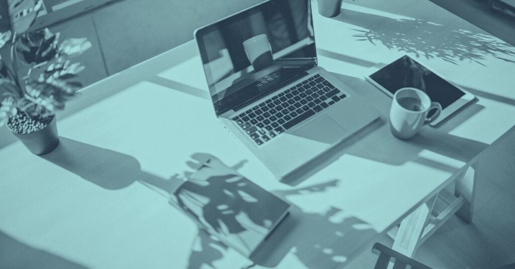 Symbolbild zum Thema Homeoffice-Betriebsstätte: Blau eingefärbte Aufnahme eines aufgeklappten Laptops auf einem Holzschreibtisch, umgeben von einer Kaffeetasse, einem Tablet, einer Grünpflanze und einem Notizbuch, mit Schattenwurf von Blättern auf der Tischplatte – repräsentative Darstellung eines typischen Heimarbeitsplatzes im Kontext von grenzüberschreitendem Homeoffice