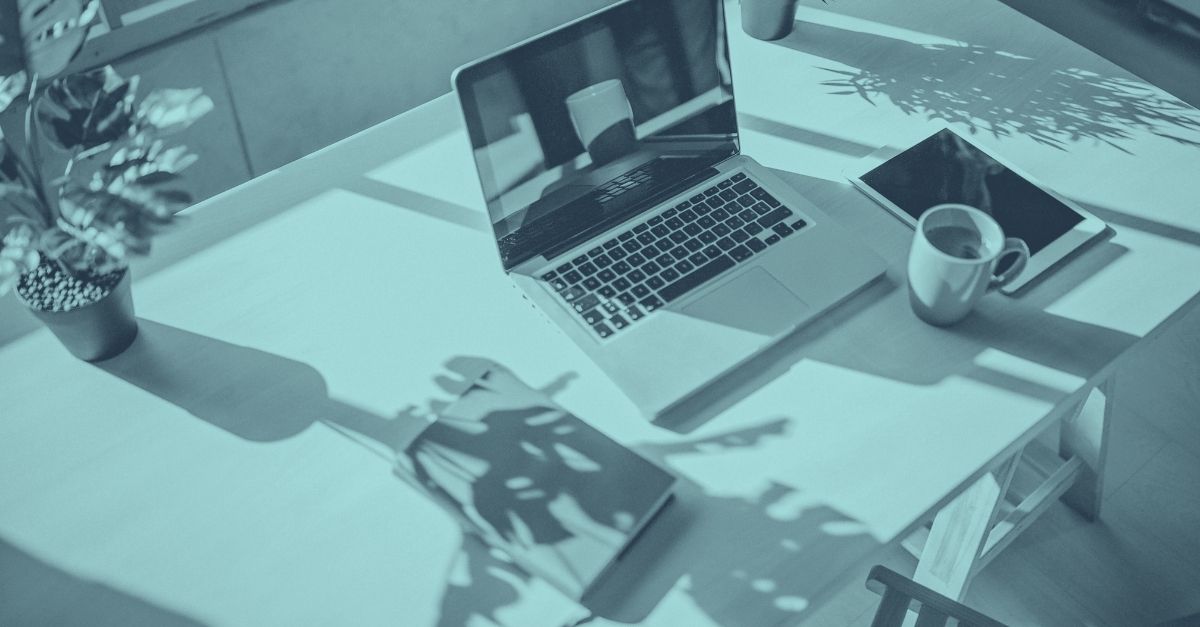 Symbolbild zum Thema Homeoffice-Betriebsstätte: Blau eingefärbte Aufnahme eines aufgeklappten Laptops auf einem Holzschreibtisch, umgeben von einer Kaffeetasse, einem Tablet, einer Grünpflanze und einem Notizbuch, mit Schattenwurf von Blättern auf der Tischplatte – repräsentative Darstellung eines typischen Heimarbeitsplatzes im Kontext von grenzüberschreitendem Homeoffice